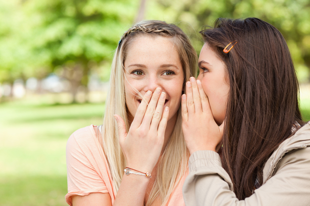 Close-up of teenagers sharing a secret with hands in front of the mouth in a park
