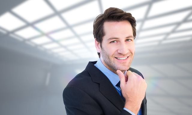 Businessman touching his chin while smiling at camera against white room with windows at ceiling